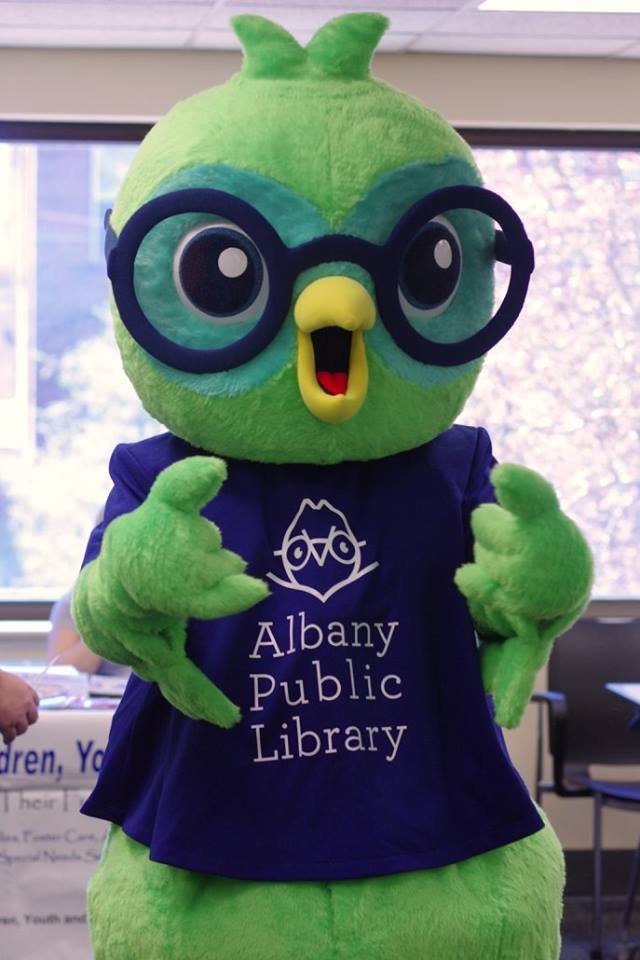 A person wearing a costume that looks like an owl with glasses wearing blue t-shirt that says "Albany Public Library"