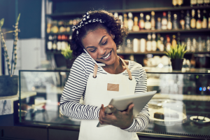 Smiling entrepreneur standing in front of the counter of her cafe talking on a cellphone and using a tablet